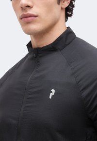 Νεαρός άνδρας που φοράει μαύρο μπουφάν με φερμουάρ και ψηλό γιακά, με μικρό λευκό γεωμετρικό λογότυπο στο αριστερό στήθος.