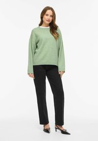 Gaisvi zaļš pārizmēra džemperis ar apaļu kaklu, kombinēts ar melnām taisnstūrveida biksēm un melnām augstpapēžu kurpēm. Mīksta tekstūra.