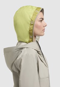 Lightweight beige jacket with a vibrant green hood. Features include a smooth texture, adjustable hood, and embroidered branding along the edge.