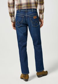 Vaqueros de mezclilla azul oscuro con corte recto, que presentan bolsillos traseros decorativos y una parche de cuero etiquetado como "Wrangler."