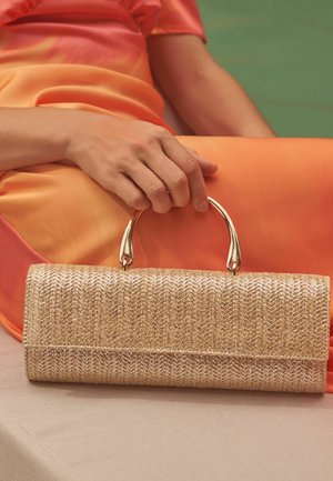 Femeie în rochie portocalie ținând o geantă plic bej texturată cu mânere aurii, așezată pe o suprafață de țesătură bej.