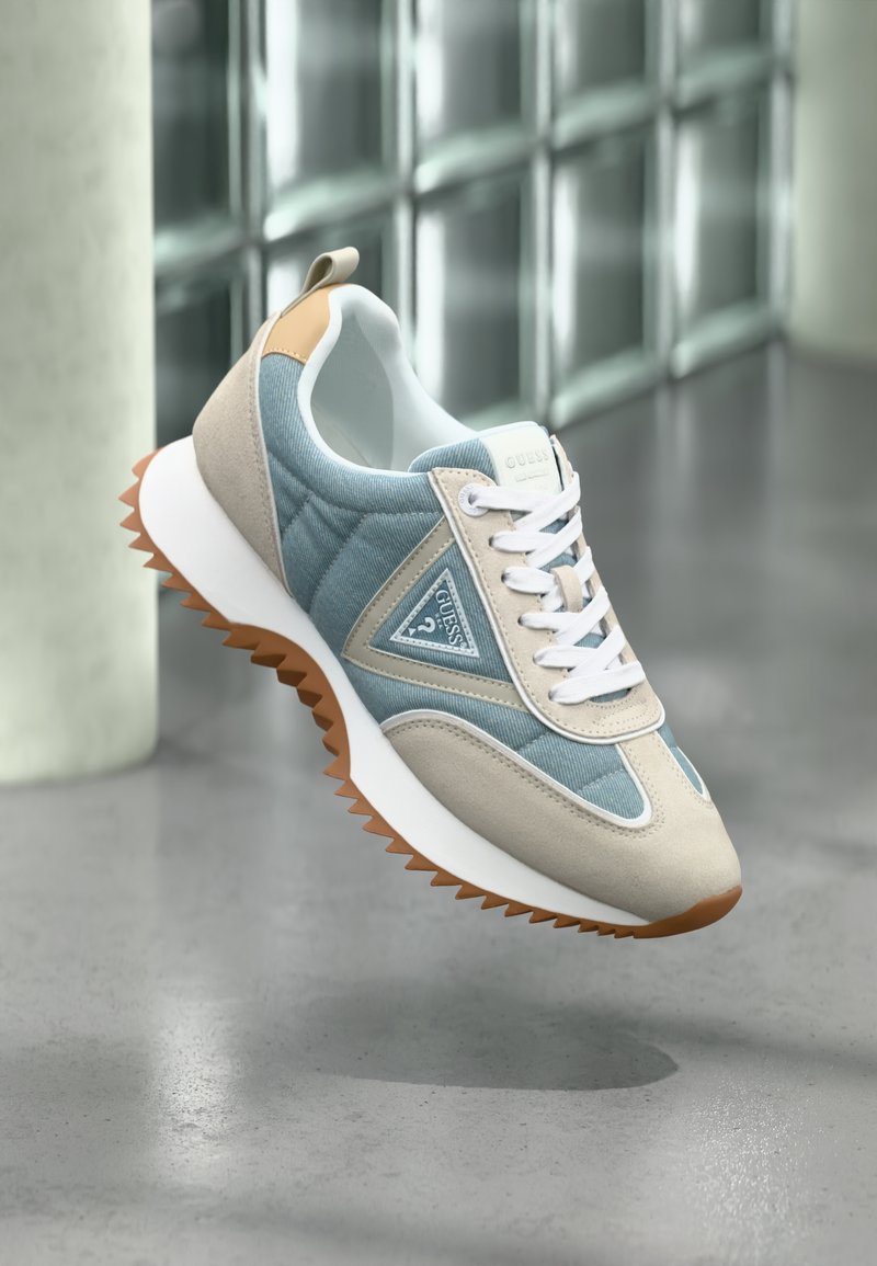 Baskets Guess beige et bleues avec des lacets blancs et une semelle brune rugueuse, présentées en apesanteur sur un fond de blocs de verre réfléchissants.
