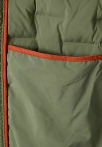 Grüner Stoff mit glatter Textur hat eine orangefarbene Tasche. Das Design ist faltbar, mit minimalen Nähten und einem leichten Erscheinungsbild.