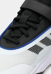 Ténis atléticos a preto e branco com um upper texturizado, pormenores em azul e atacadores pretos. Detalhes notáveis incluem uma tira e um design riscado.