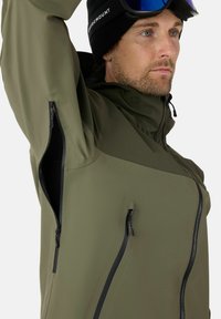 Giacca impermeabile verde oliva con colletto alto, tasche con zip e texture liscia. Presenta cerniere nere e un design minimalista.