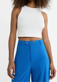 Weißes geripptes Crop-Top mit hohem Kragen, kombiniert mit leuchtend blauen Hosen, die einen Knopfverschluss und eine glatte Textur aufweisen.