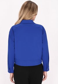 Jachetă albastră scurtă dintr-un material neted, cu guler, manșete cu nasturi și un design ajustat, purtată deasupra unor pantaloni negri.