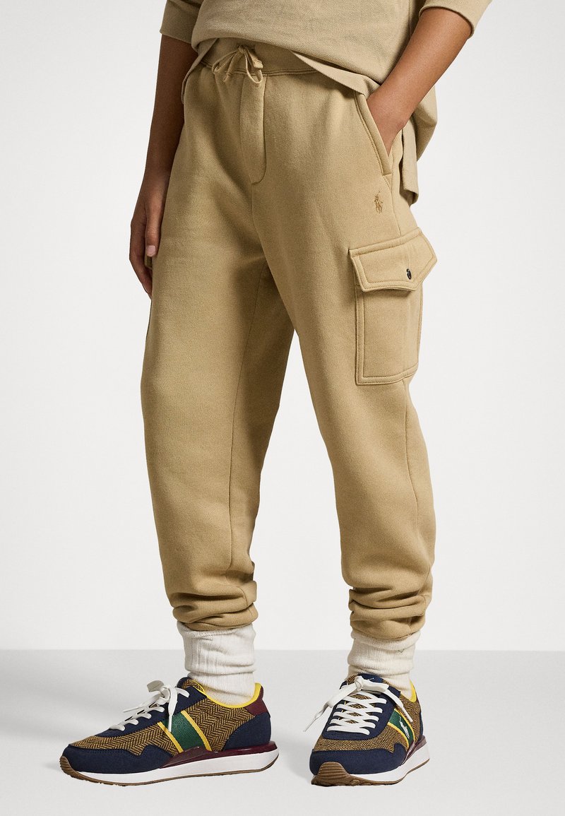 Calças de moletão bege com punhos elásticos, cordão na cintura e bolso cargo no lado esquerdo. Combinadas com ténis multicoloridos e meias brancas até ao tornozelo.