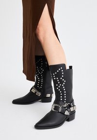 Botas negras hasta el tobillo con punta afilada, adornadas con tachuelas y cadenas plateadas, con cierre de cremallera y un tacón bajo y ancho.