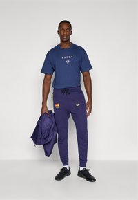 Marineblå t-shirt med "BARCA" print, parret med lilla træningsbukser med Nike-logo og FC Barcelona-emblem. Sorte sneakers.