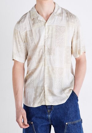 Man wearing beige patterned short-sleeve button-up shirt with blue denim shorts, one hand in pocket against white background.
