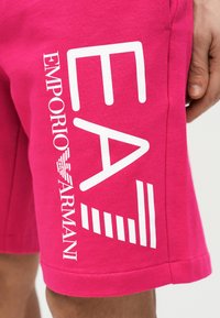 Φωτεινά ροζ σορτς με μεγάλο λευκό λογότυπο "EA7 Emporio Armani" στον μηρό, φορεμένα από ένα άτομο που έχει το δεξί του χέρι κοντά.