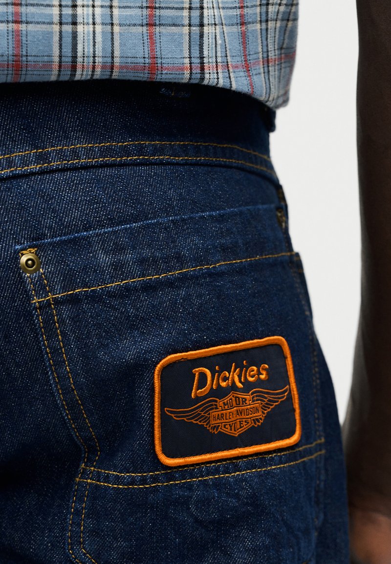 Gesäßtasche einer Jeans aus Denim mit einem aufgenähten Patch mit orange umrandetem Dickies- und Harley-Davidson-Logo unter einem in die Hose gesteckten karierten Hemd.