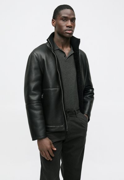 Črna usnjena jakna z visokim ovratnikom, zadrgo in kontrastno mehko podlogo. Kombinirana s temnim polo majico in črnimi hlačami.