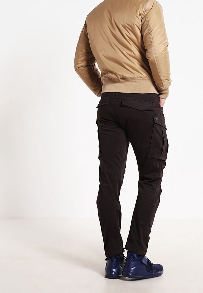 Homme portant une veste bomber beige, un pantalon cargo noir et des baskets bleu foncé, debout les mains dans les poches, dos tourné.