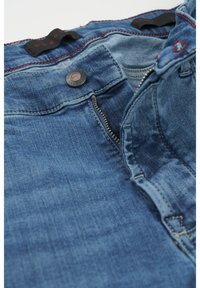 Dżinsy w jasnoniebieskim odcieniu z zapięciem na suwak, zapięciem na guzik oraz kontrastującymi czerwonymi stymi. Zawierają etykietę materiałową umieszczoną wewnątrz pasa.