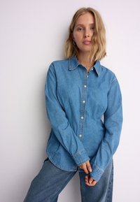 Lichtblauw denim overhemd met een knoopsluiting aan de voorkant, lange mouwen en een klassieke kraag, voorzien van subtiele stiksels en een losse pasvorm.