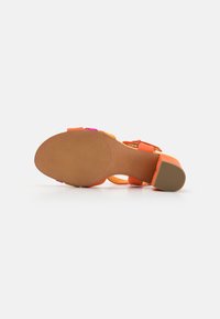 Sandale en daim orange avec une bride de cheville, un talon bloc et une semelle extérieure en caoutchouc texturé. Présente des accents rose et violet sur les brides.