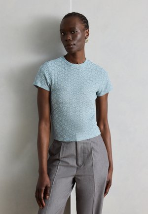 Femme aux cheveux tressés portant un haut à manches courtes texturé bleu clair et un pantalon gris à taille haute sur un fond uni.