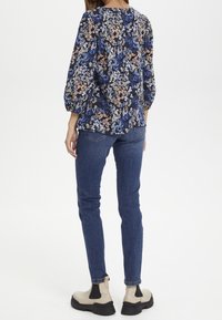 Blouse florale bleu foncé avec des manches bouffantes, associée à un jean foncé ajusté et des bottines beiges avec des panneaux latéraux noirs.