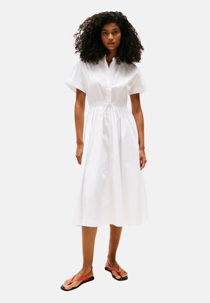 Weißes Baumwollhemdkleid mit kurzen Ärmeln, Knopfleiste und tailliertem Schnitt. Model trägt rote Flip-Flops und steht vor einem schlichten Hintergrund.