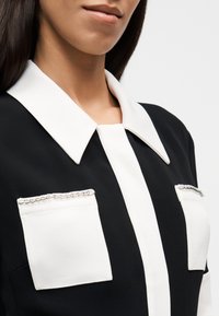 Γυναίκα που φοράει μαύρο ένδυμα με μεγάλο λευκό γιακά και δύο λευκές τσέπες στο στήθος, κάθε μία διακοσμημένη με ασημένιο αλυσίδα περιθώριο.