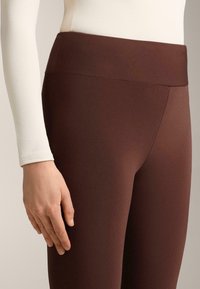 Rjave legice visokega pasu, izdelane iz gladkega, raztegljivega blaga. Oblikovanje vključuje brezšiven pas in prilegajoč silhueto.