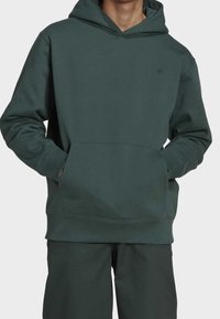 Man i mörkgrön luvtröja med framficka och matchande mörkgröna byxor, händerna i fickorna, mot en enfärgad bakgrund.