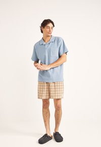 Polo de manga corta azul claro con un bolsillo, combinado con pantalones cortos de cuadros beige. El modelo completa el atuendo con zapatos oscuros tipo slip-on.
