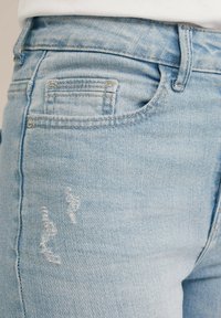 Svetlo modri trendi jeans s obdarjenim krojem, petimi žepe in poškodovanim detajlom z raztrganimi luknjami spredaj za teksturo.