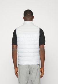 Ljusgrå pufferväst med hög krage och horisontell kviltad design, bärs över en svart kortärmad skjorta. Slät textur och minimal detaljering.