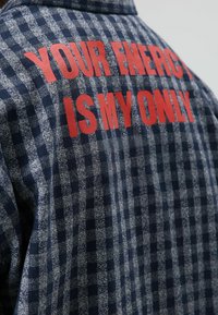 Marineblaue und graue kariertes Flanellhemd mit rotem Text auf der Rückseite: "DEINE ENERGIE IST MEINE EINZIGE." Weicher, strukturierter Stoff, sichtbare Nähte.