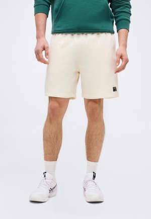 Homme portant un short crème mi-mollet, des chaussettes de sport blanches, des baskets blanches et un haut vert à manches longues, debout devant un fond uni.