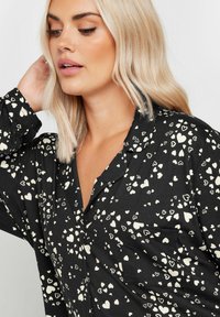 Camisa negra con cuello, con un patrón de corazones blancos dispersos, mangas largas y un pequeño bolsillo en el pecho. Textura de tela suave.