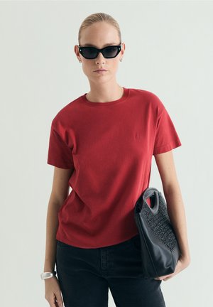 Donna con t-shirt rossa e occhiali da sole neri, che tiene una borsa nera con texture su uno sfondo chiaro e uniforme.