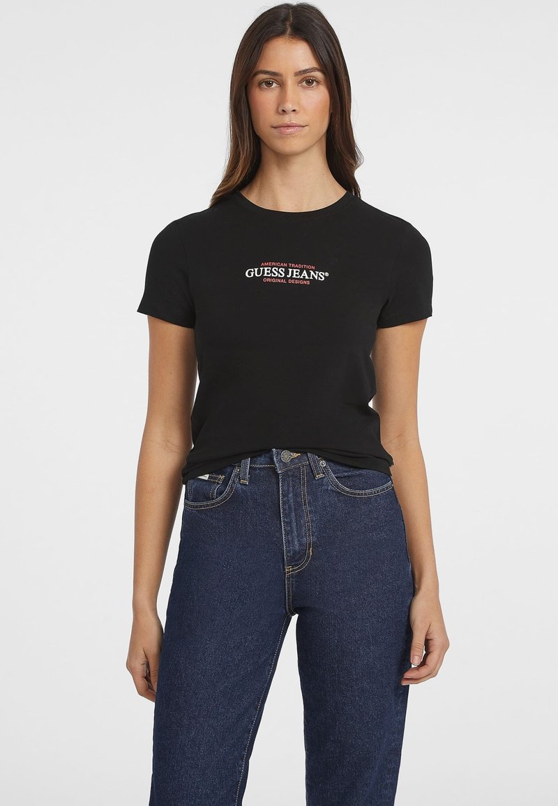 Čierne bavlnené tričko s okrúhlym výstrihom a krátkymi rukávmi, zdobené vyšívaným logom "GUESS JEANS" v bielej a červenej farbe. Nosené s modrými džínsami s vysokým pásom.