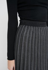 Falda plisada negra con brillo metálico, que presenta una cintura con textura grabada con "KARL LAGERFELD." Las rayas verticales enfatizan el diseño.