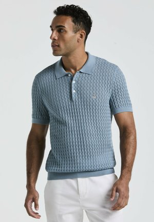 Man in een lichtblauw gestructureerd poloshirt met korte mouwen en witte broek, kijkt naar links tegen een effen lichte achtergrond.