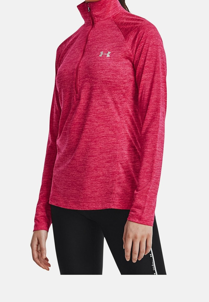 Kvinna iklädd en röd långärmad tröja med halv dragkedja för sport och svarta leggings står mot en enkel ljus bakgrund.