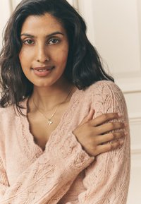 Jasnoróżowy sweter na drutach z misternymi wzorami warkoczy, z dekoltem w kształcie litery V. Widoczny jest delikatny złoty naszyjnik z małym wisiorkiem.