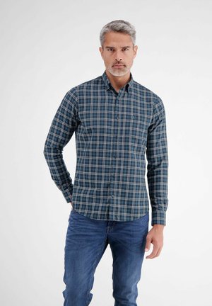 Mann mit grauen Haaren, der ein blau-grün kariertes Hemd mit Knöpfen und blaue Jeans trägt und vor einem einfarbigen weißen Hintergrund steht.