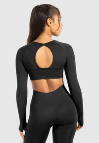 Kobieta w czarnej, długiej bluzce crop top z owalnym wycięciem na plecach i dopasowanych, wysokich legginsach, stojąca na tle jasnoszarym.