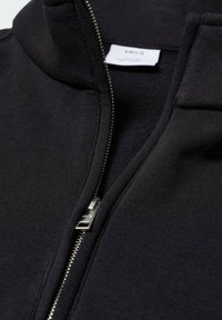 Giacca nera con zip realizzata in tessuto morbido, con collo alto e cerniera argentata. Presenta un'etichetta interna con le informazioni del marchio.