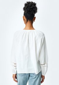 Hvid bluse med lange, bløde ærmer. Har blonde detaljer ved manchetterne og en sammensnøret halsudskæring, lavet af letvægtsstof.