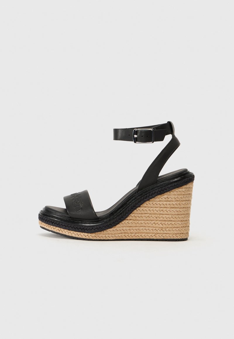 Black leather wedge sandals with a strap and buckle, featuring a jute and black rope-wrapped heel, smooth texture, and logo detail.