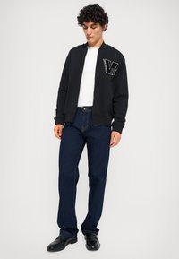 Črna zip-up dukserica z rebrastimi zapestji, ki ima belo vezeno logotip na prsih. Nosena nad belo majico in temno modrimi kavbojkami.
