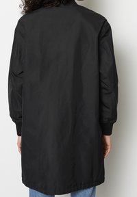 Persona con una larga chaqueta bomber negra con puños acanalados, visible desde la espalda, combinada con jeans azules, frente a un fondo sencillo.