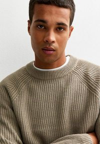 Pull en tricot brun côtelé avec un col rond et des manches raglan, porté sur un t-shirt blanc, tissu texturé avec des coutures apparentes.