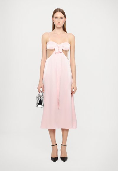 Vestido de satén rosa con un cuerpo ajustado y detalle de lazo, que fluye hasta una longitud midi. Presenta tirantes finos y laterales descubiertos. Bolso plateado y tacones negros.