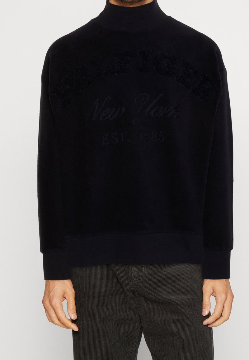 Granatowa bluza polarowa z wysokim kołnierzem, z haftowanym napisem „HILFIGER” i „New York”. Miękka w dotyku, o swobodnym kroju, z ściągaczami na mankietach.